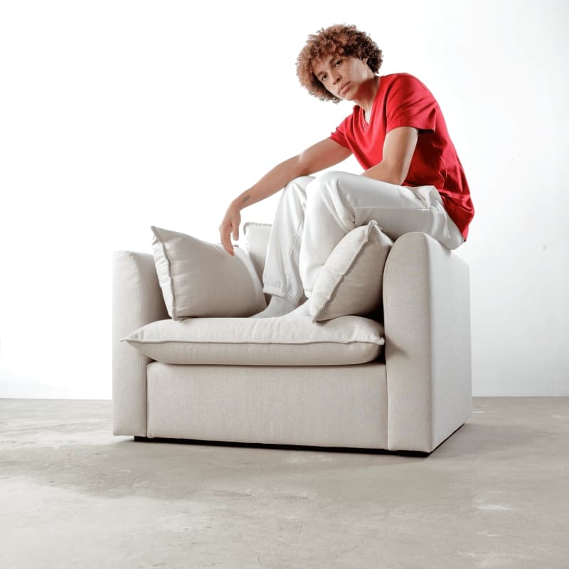 A young man in a red t-shirt sits on the back edge of a beige Scooch Daisy armchair
