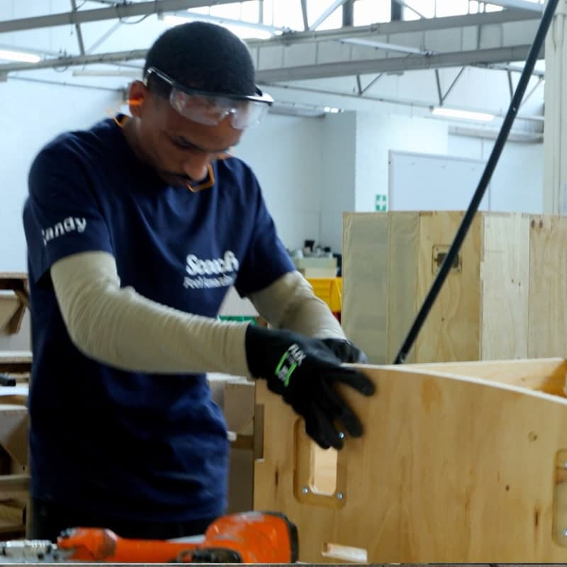 A Scooch factory worker, in a blue Scooch-branded t-shirt, works to assemble a Scooch sofa frame in the Scooch factory